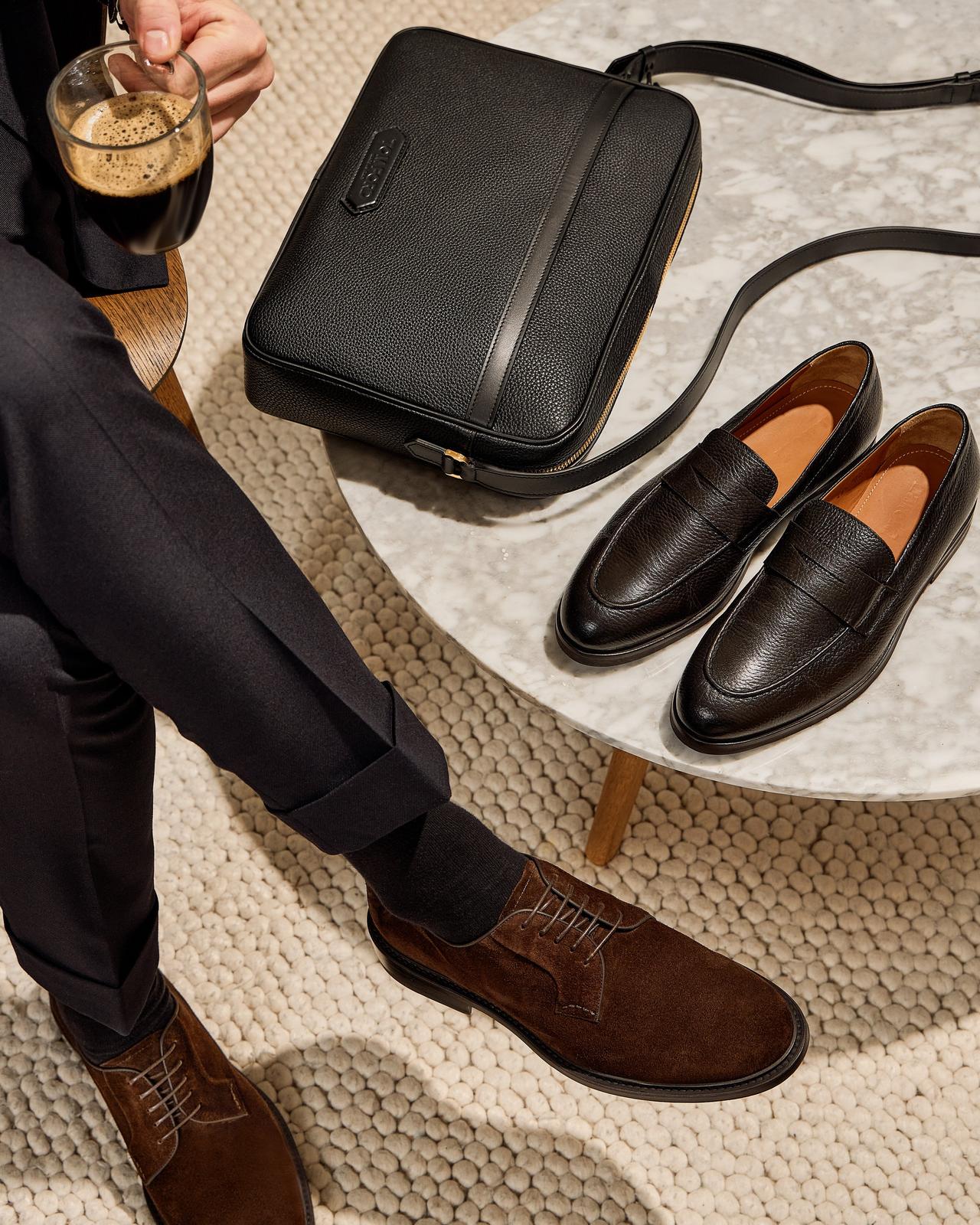 A man sitting with coffee beside a table with shoes and a bag on it
