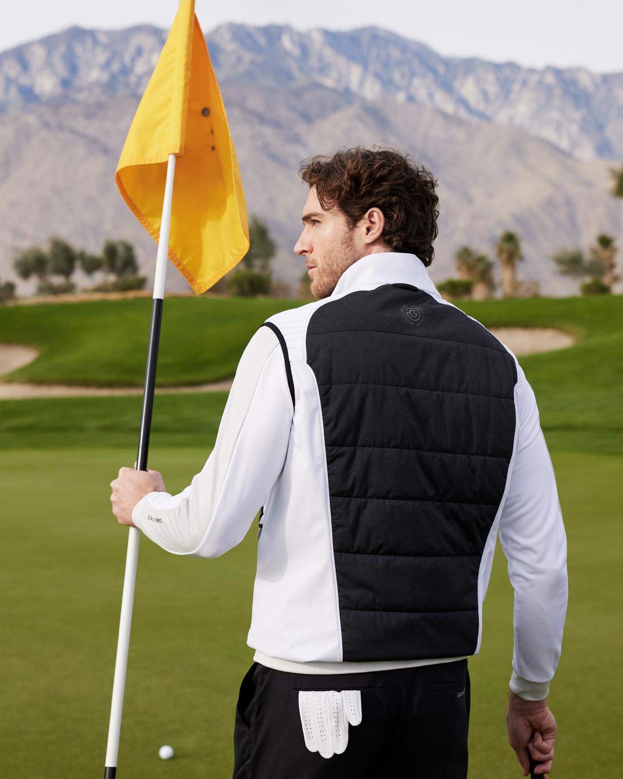 male golfer facing back, holding a flag, wearing a vest, sweater and shorts