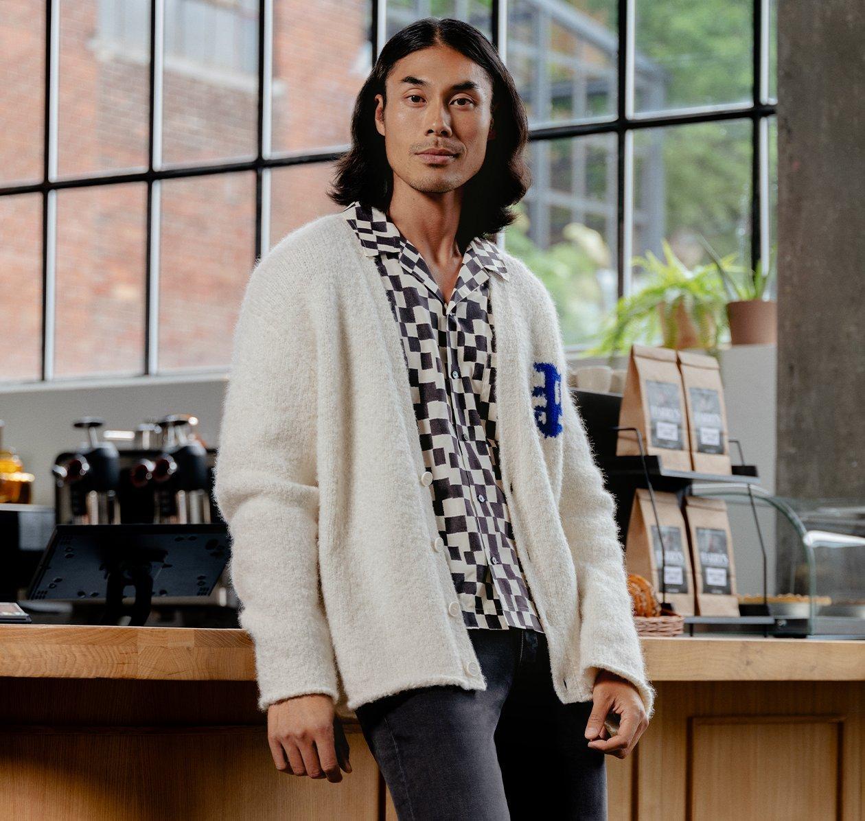 A model in a checkered shirt and sweater leaning against a counter