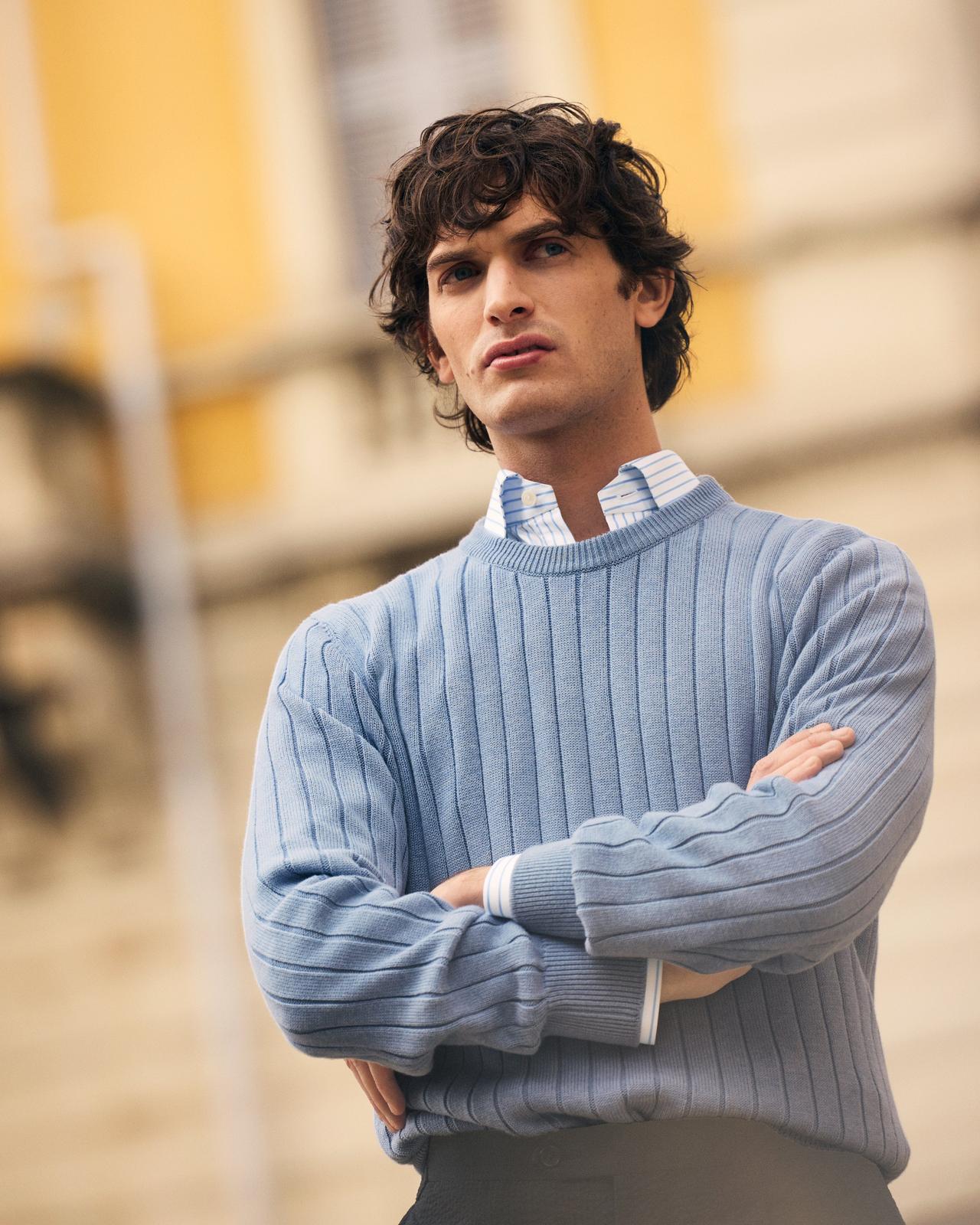 A man wearing a light blue grey sweater with white and blue striped shirt