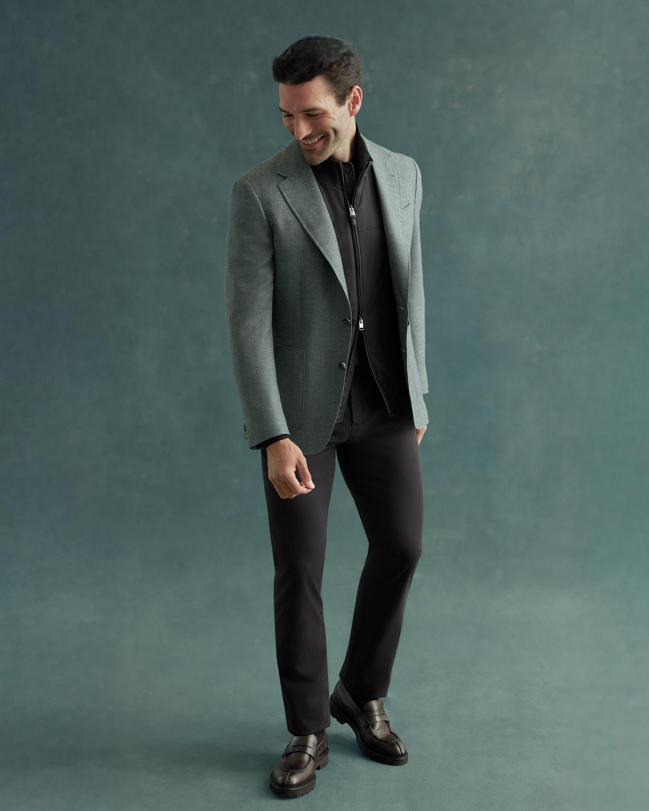 A man wearing a green Canali blazer over a black shirt and black pants with black dress shoes