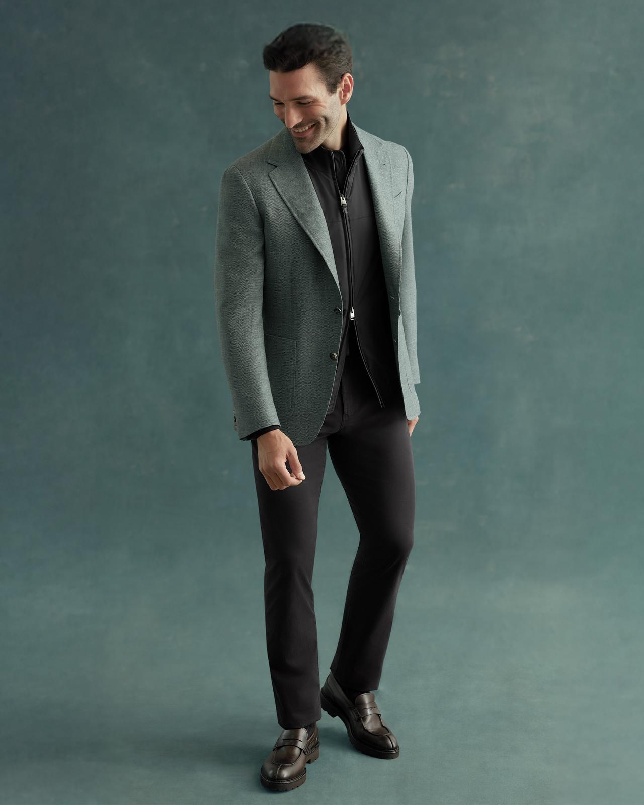 A man wearing a green Canali blazer over a black shirt and black pants with black dress shoes
