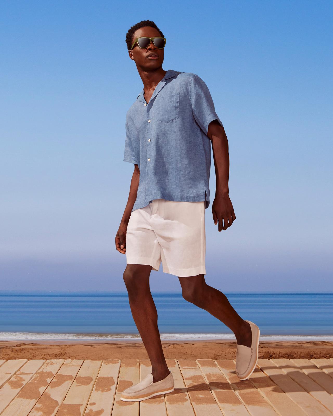 A man in sunglasses and a blue shirt walking on a beach.