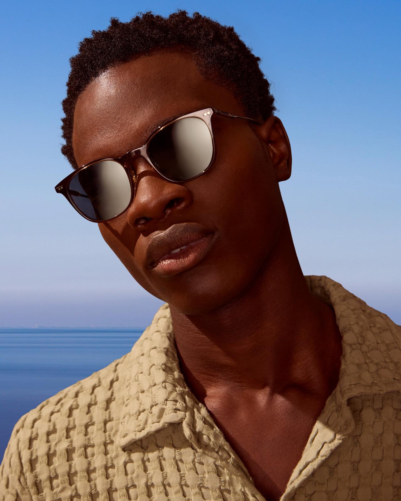 A man in sunglasses standing in front of the ocean with a blue sky.