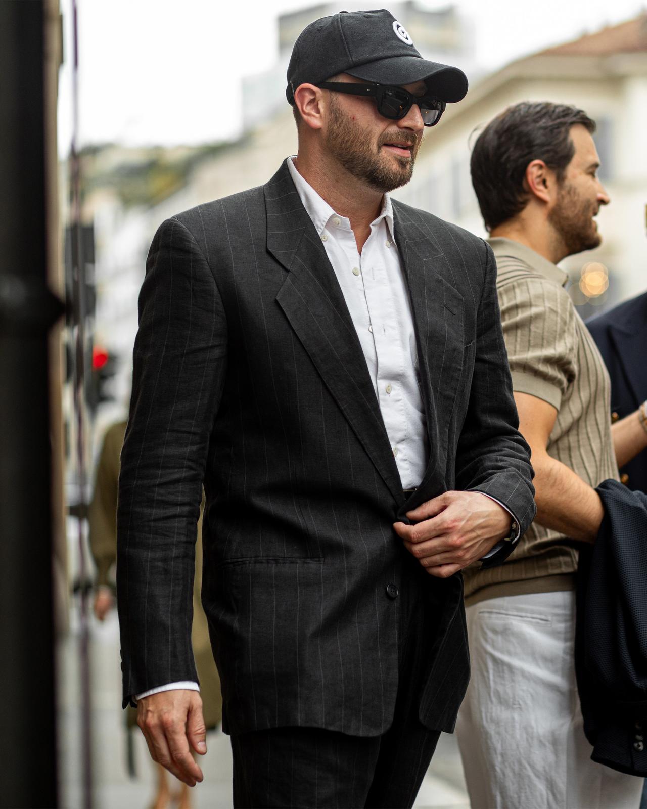 homme à l'extérieur portant un costume à fines rayures, une chemise blanche, une casquette de baseball et des lunettes de soleil