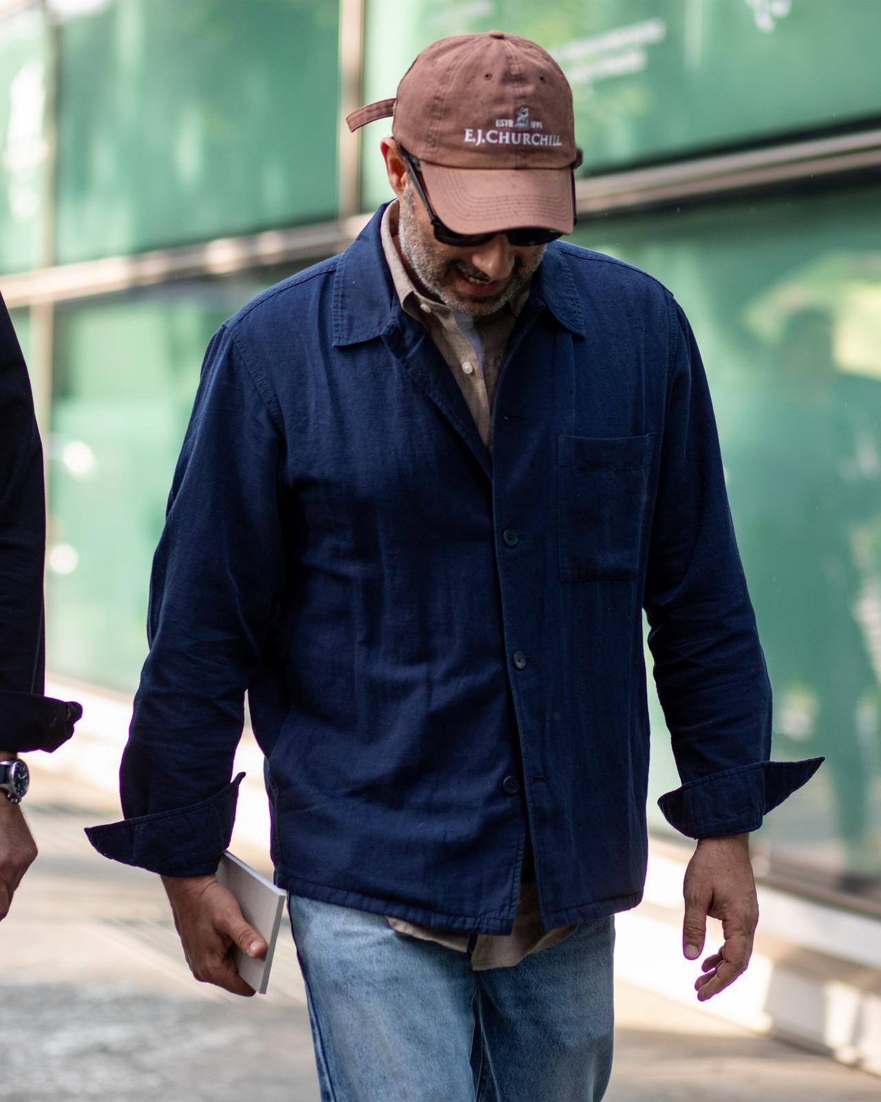 man walking outside with head down wearing sunglasses, baseball cap, overshirt and jeans holding notepad