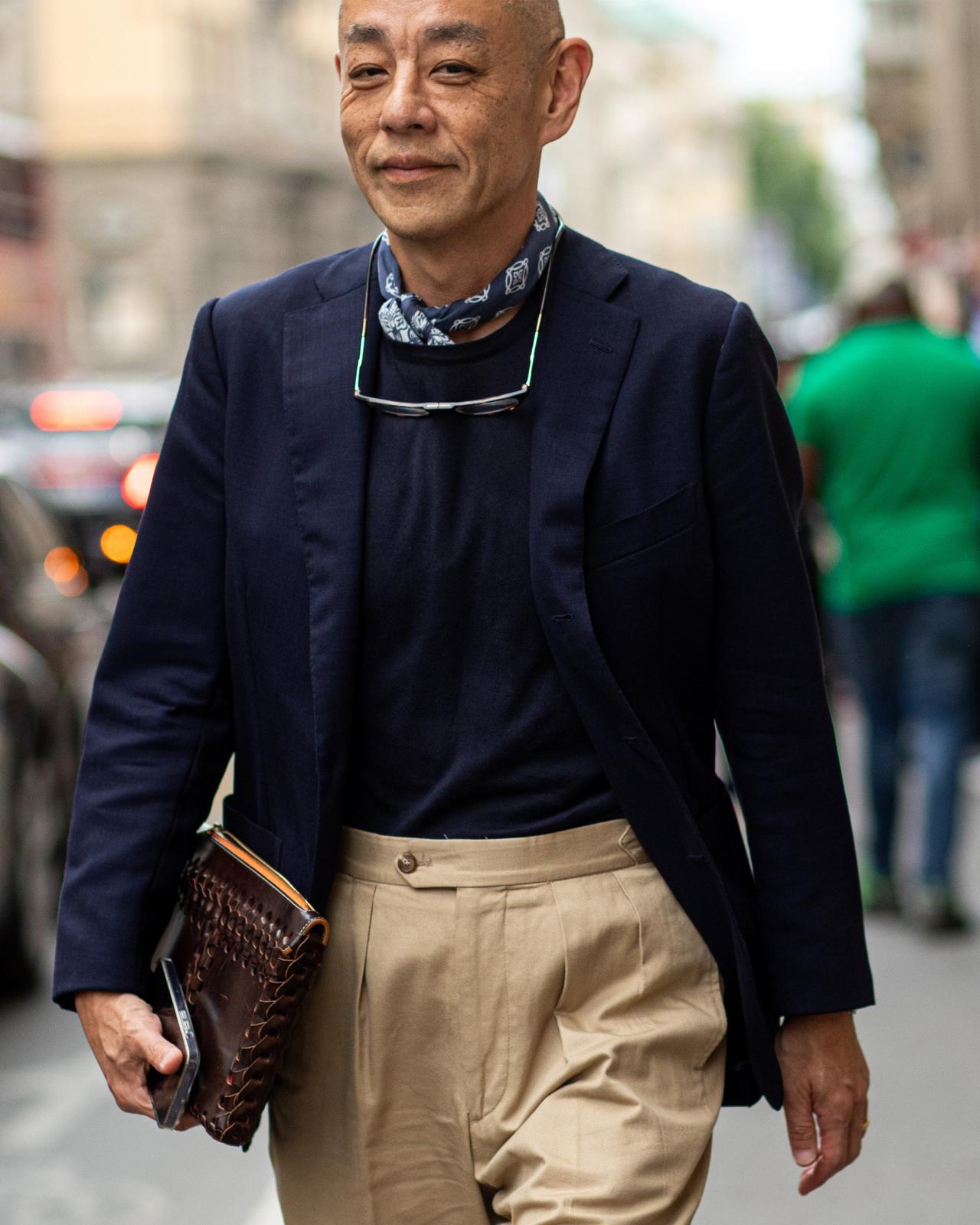 male walking in the street with neck scarf, sunglasses, sport jacket, dress pants while holding clutch