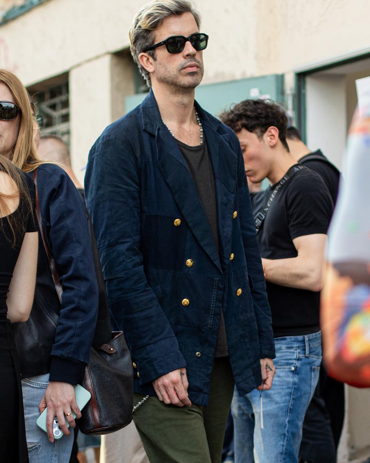 male in sunglasses walking outside wearing navy double breasted jacket and green jeans