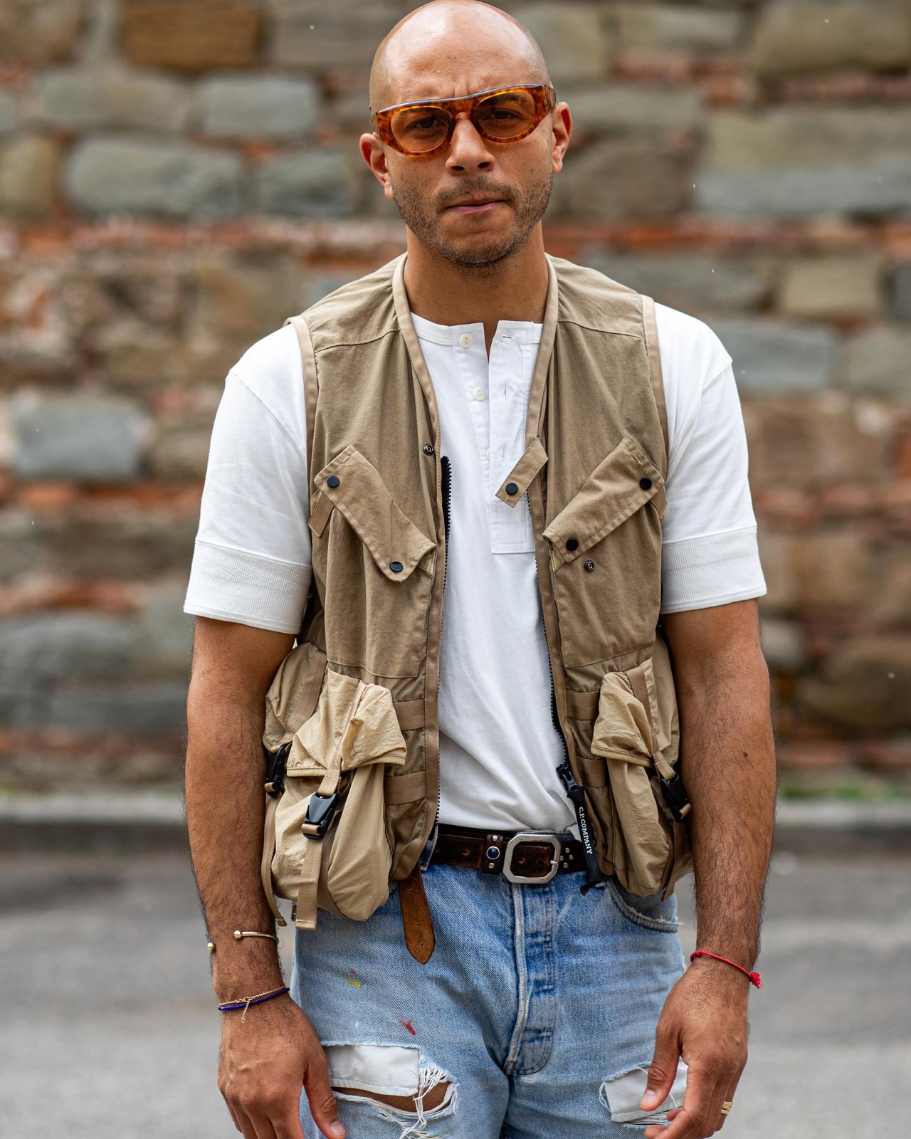 a man in a vest and jeans standing in front of a wall