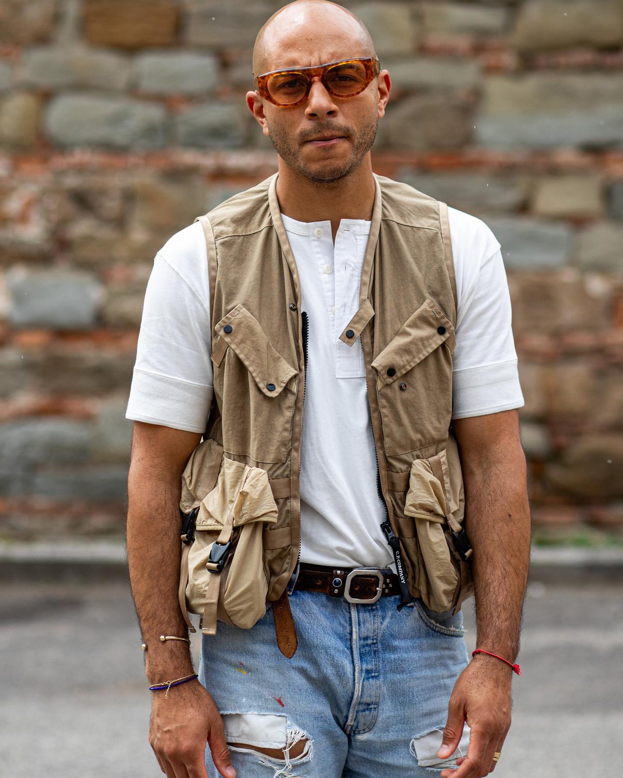 un homme dans un gilet et un jean debout devant un mur