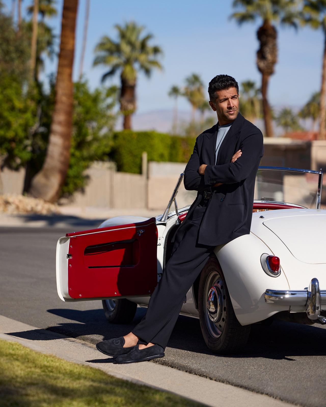 a man in a navy suit leaning against a white and red car