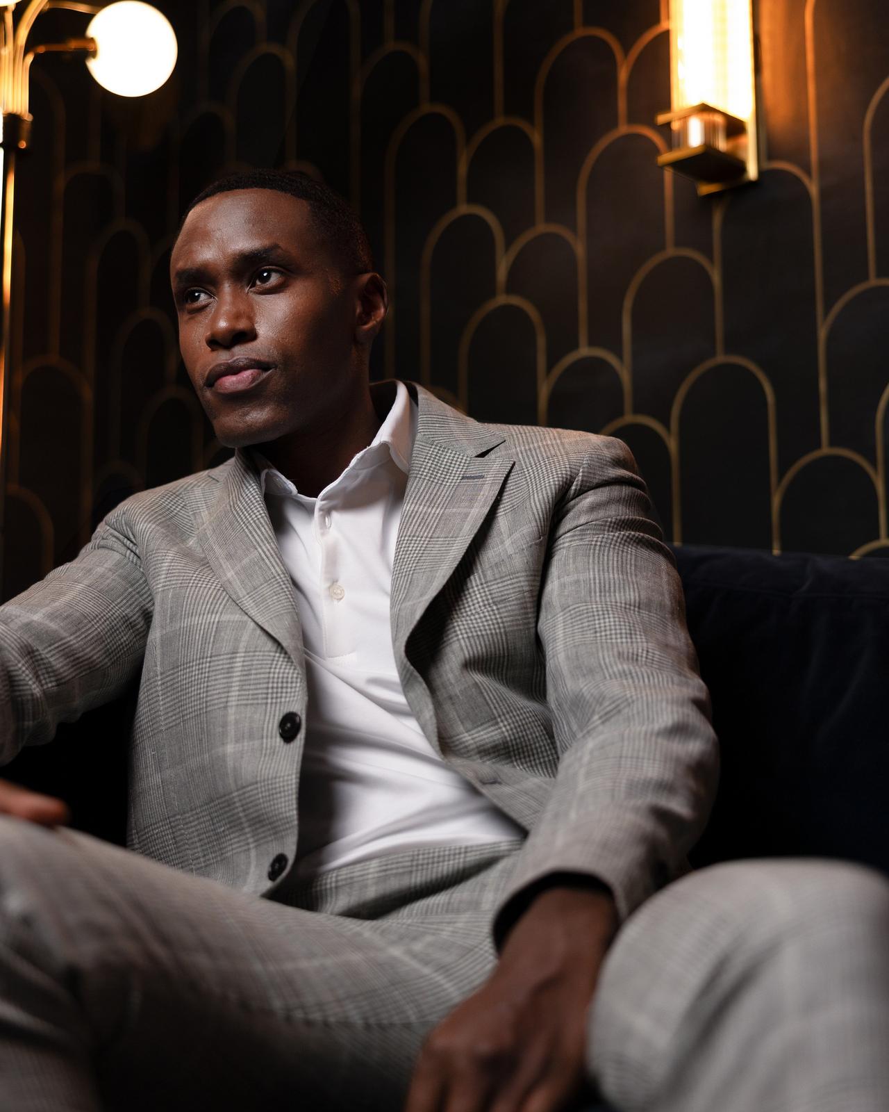 a male in a grey checked suit and white polo sitting