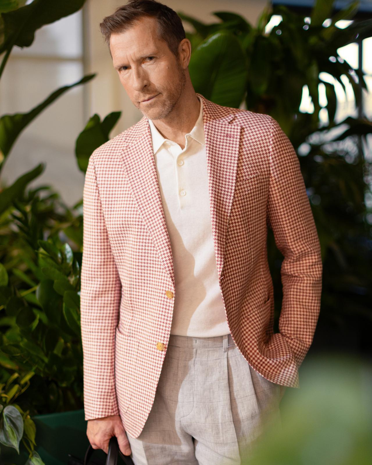 a male in a white polo and pink checked sports jacket with trees in the background