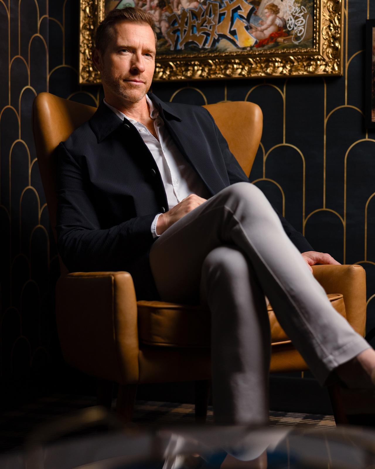 a male sitting in a camel armchair wearing a blue jacket and grey pants