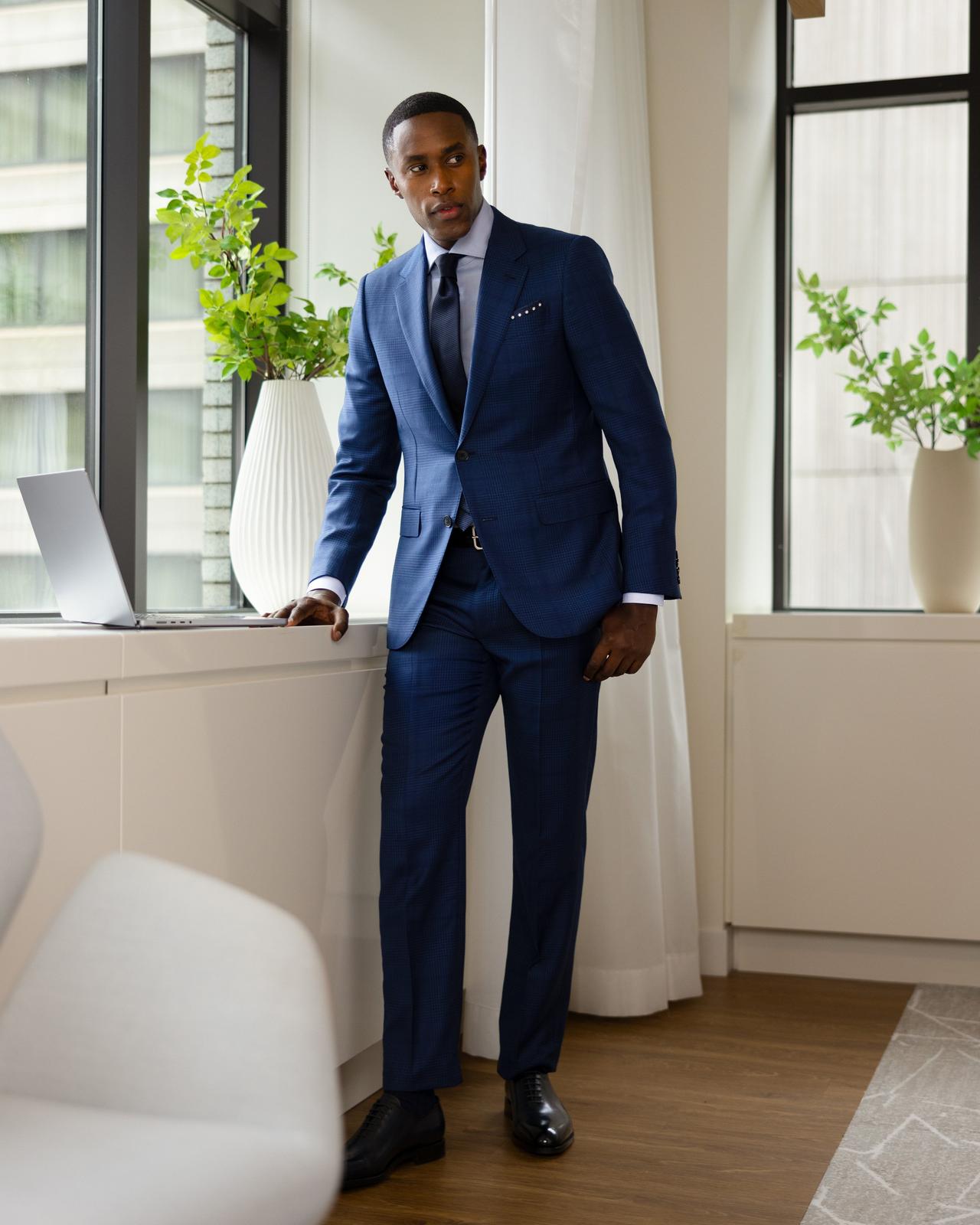 a male wearing a blue suit, shirt and tie working on a laptop near an office window