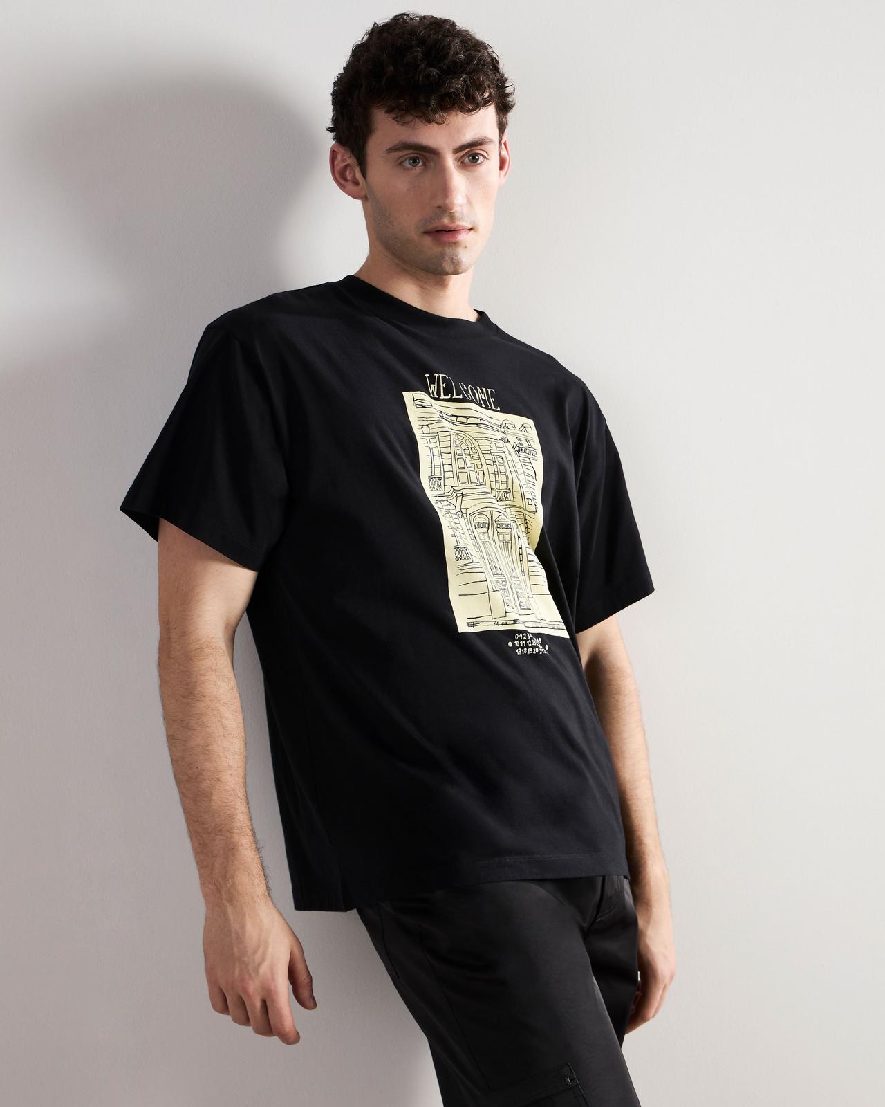 a man in black pants leaning against a wall wearing a black graphic t-shirt
