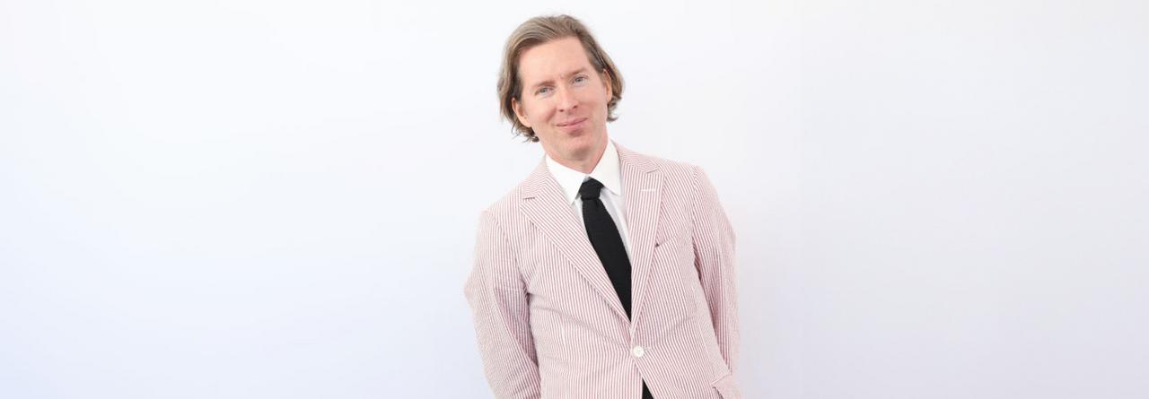Wes Anderson wearing a light pink pinstriped suit with a black tie