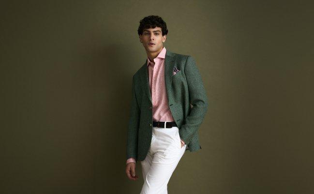 A male model wearing a forest green blazer, paired with a pink dress shirt, leather belt and white pants