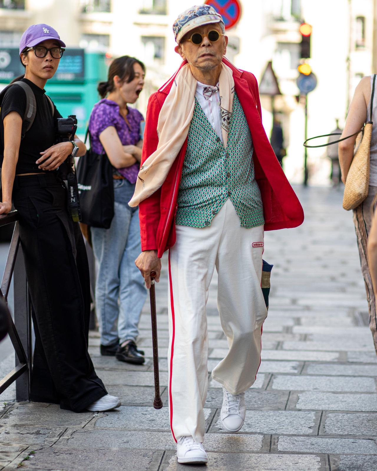 Aman walking down a street with a red sport jacket, cane and a hat