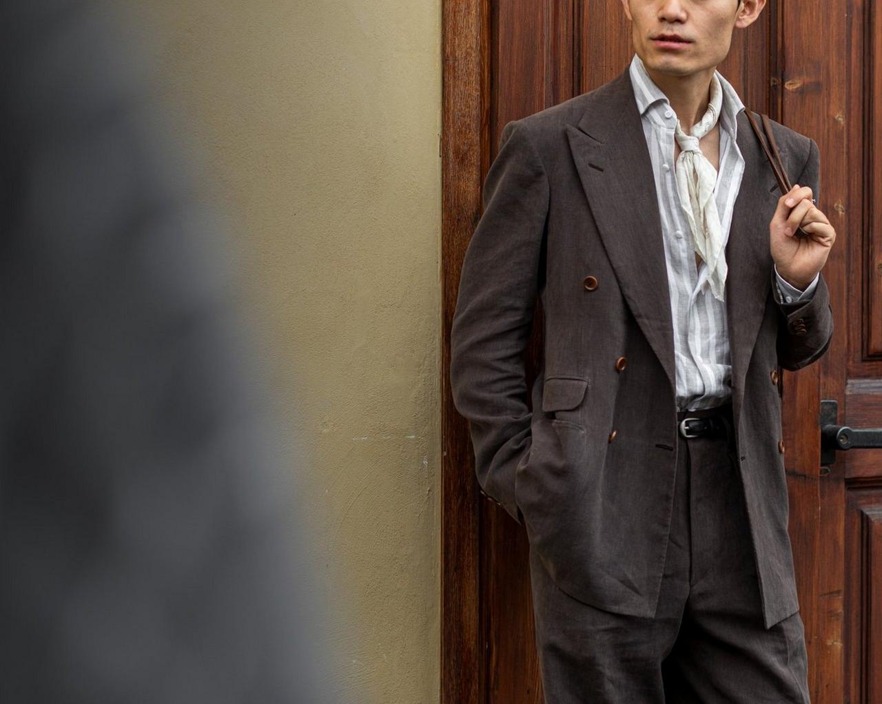 Un homme portant un manteau superposé à une chemise grise à rayures, avec un pantalon noir