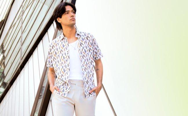 Un homme portant une chemise blanche à manches courtes à motif, un t-shirt blanc en dessous, et un pantalon beige