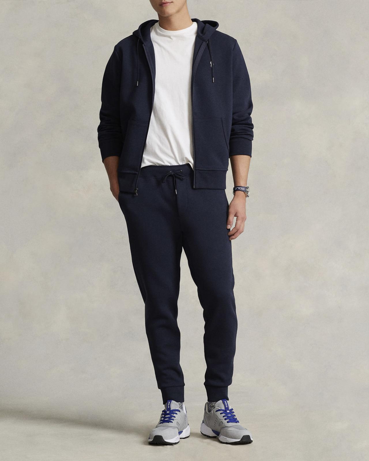 A model wearing a Polo Ralph Lauren Jogger Pant in navy paired with a matching zip-up hoodie, a white T-shirt, and gray sneakers