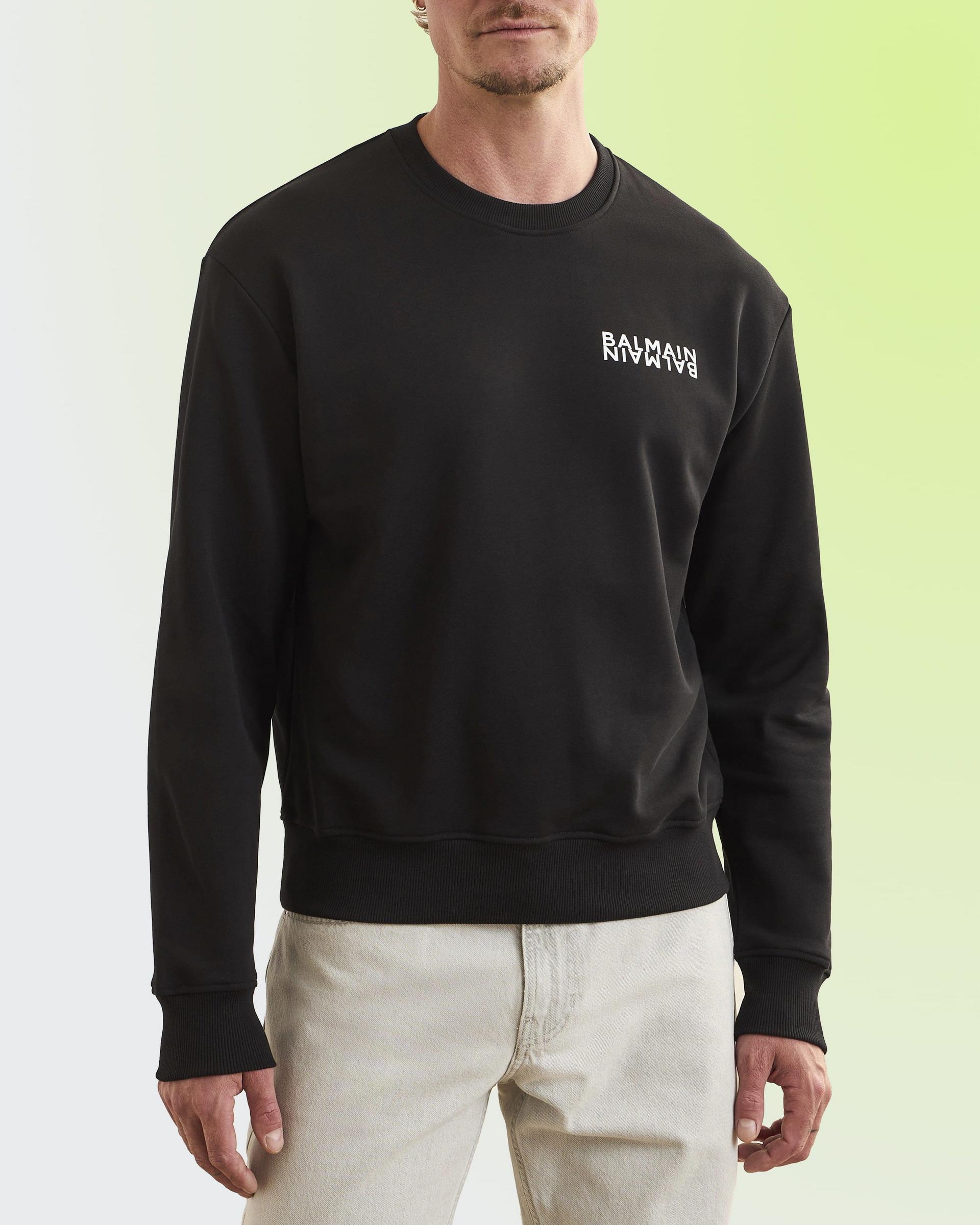 A man wearing a black crew neck sweatshirt with a small white logo on the chest, paired with light-colored pants