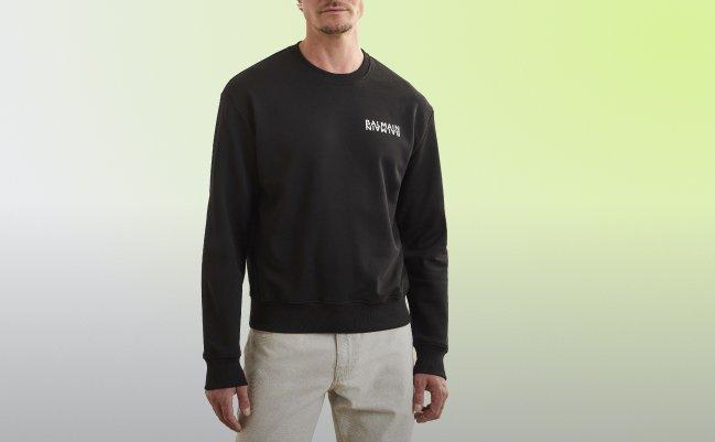 Un homme portant un sweat-shirt noir à col rond avec un petit logo blanc sur la poitrine, accompagné d’un pantalon de couleur claire
