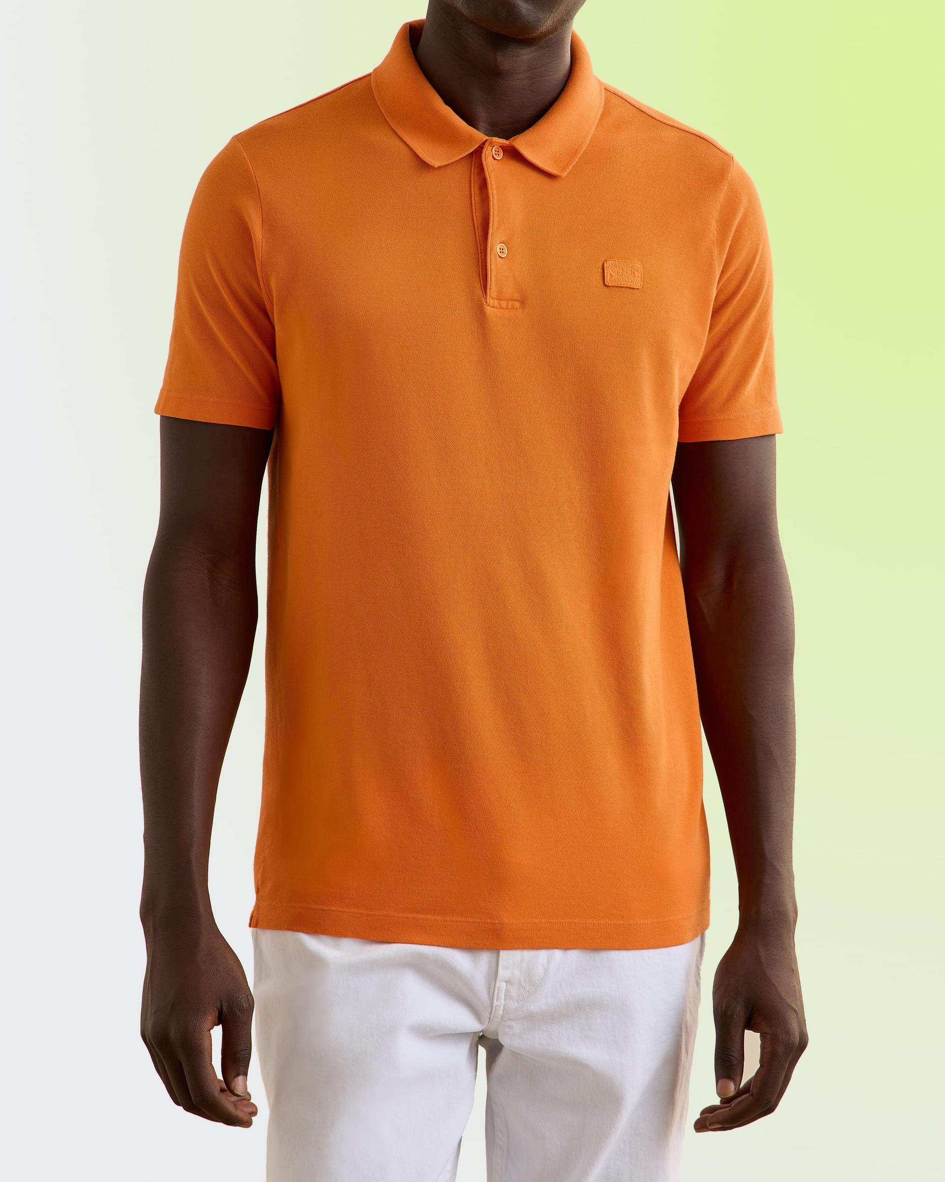 mannequin portant un polo orange vif associé à un pantalon blanc