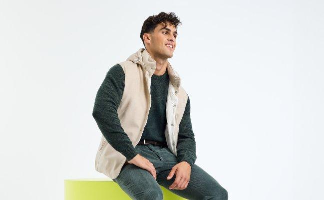 A male model wearing a beige vest with a dark grey knit sweater, styled with black denim