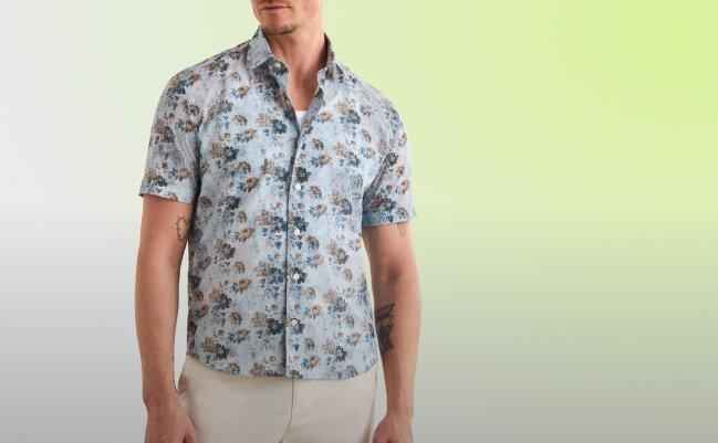 Un homme portant une chemise à manches courtes boutonnée à imprimé floral, sur fond bleu pâle avec un motif floral tropical