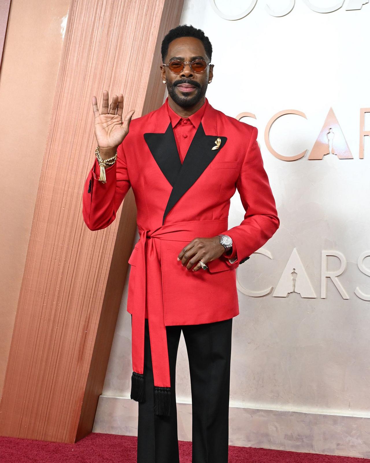 Colman Domingo wearing a red belted cocktail jacket with black pants standing on red carpet
