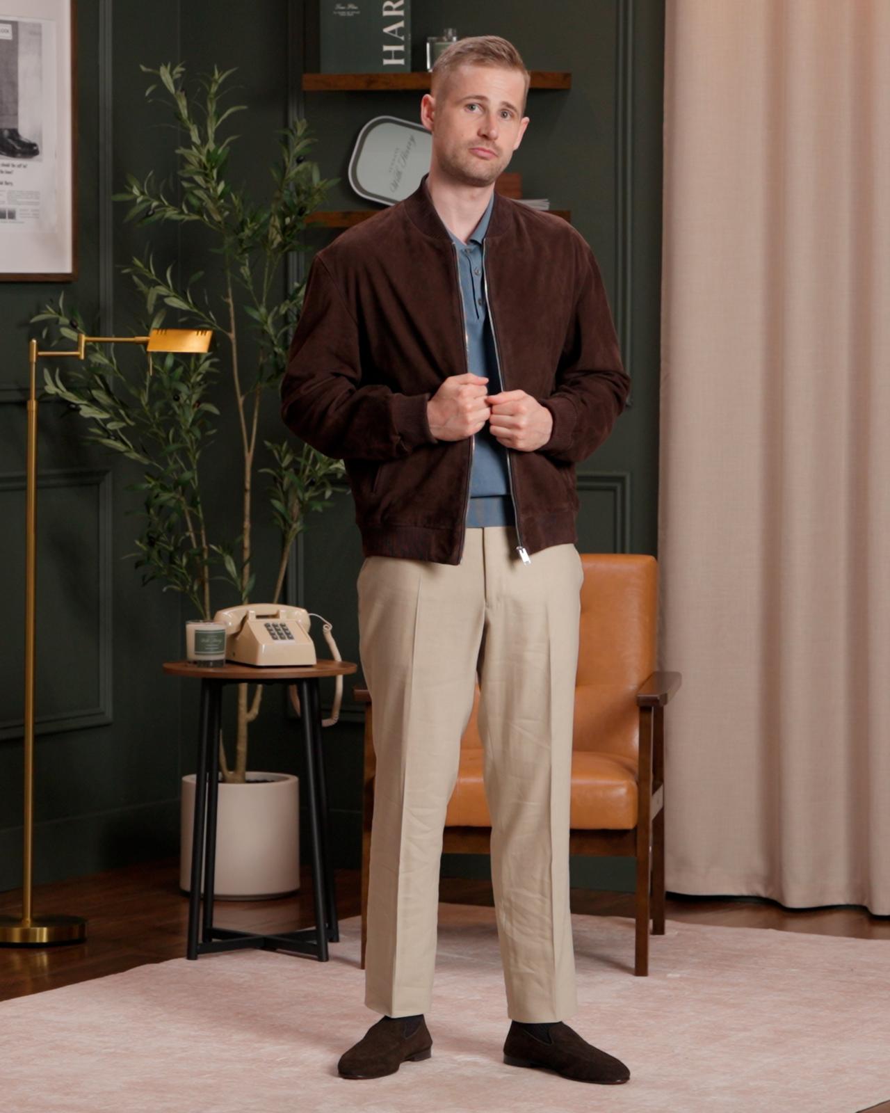 Fashion model wearing a dark brown suede jacket over a blue collared shirt, paired with beige trousers and dark brown shoes