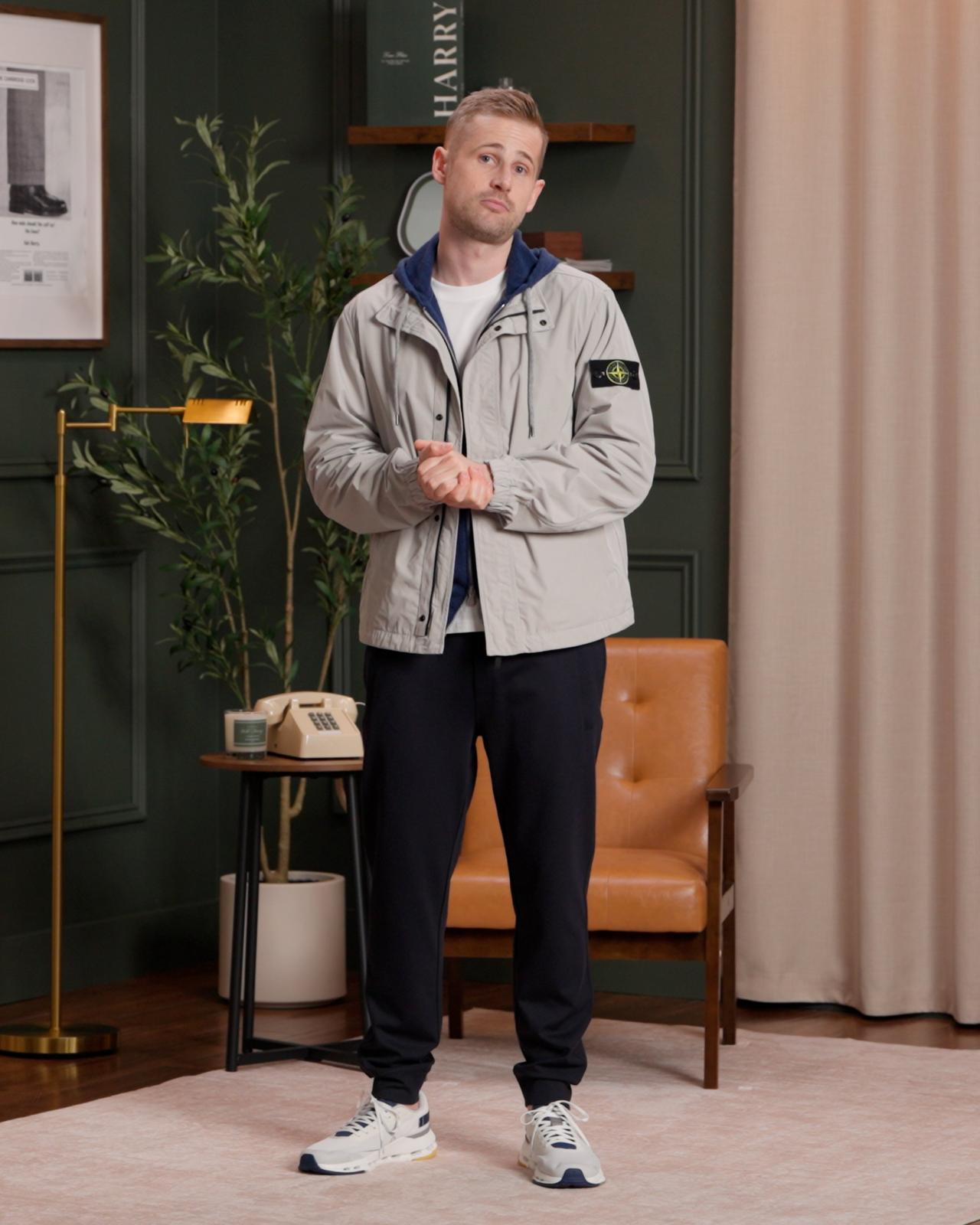 Fashion model wearing a light grey jacket over a navy blue zip-up hoodie and a white T-shirt, with hands in the hoodie pockets