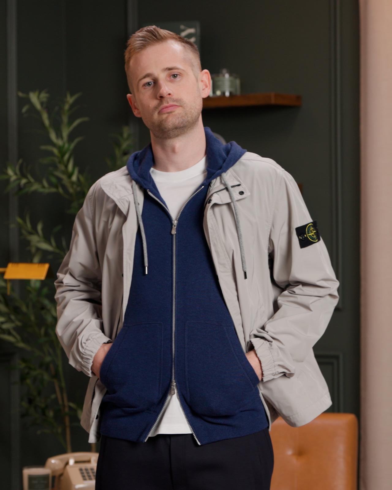 Plan rapproché d’un mannequin de mode portant une veste gris clair par-dessus un sweat à capuche zippé bleu marine et un t-shirt blanc, les mains dans les poches du sweat à capuche