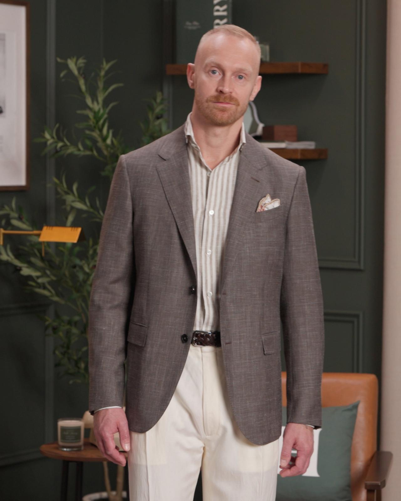 Un homme portant un manteau brun fumé, une chemise rayée blanc cassé et brun pâle, ainsi qu’un pantalon blanc cassé