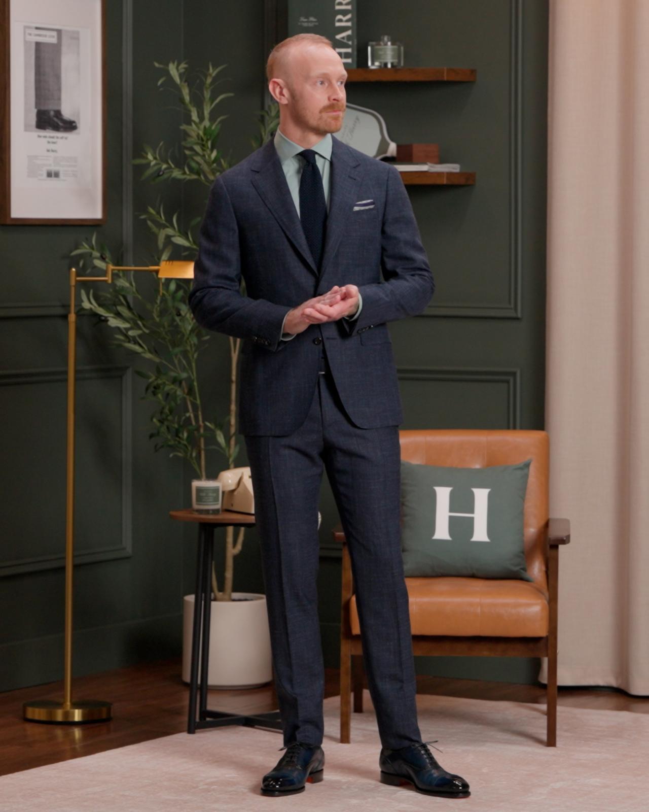 A man wearing a dark blue-gry color suit with dusty mauve shirt, dark blue tie and light blue pocket square