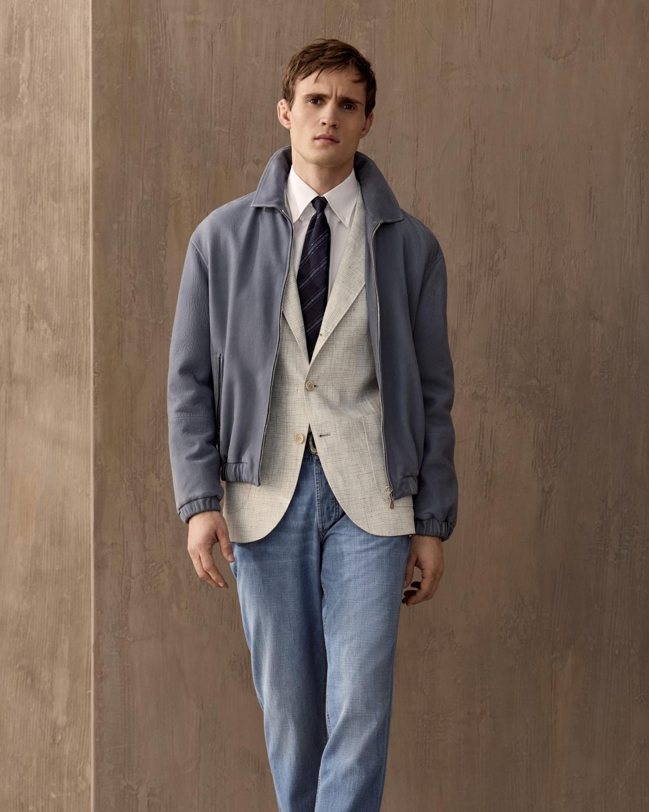 A model wearing a slate grey lightweight Brunello Cucinelli jacket, paired with an off white sport jacket, white dress shirt, black tie, and blue denim