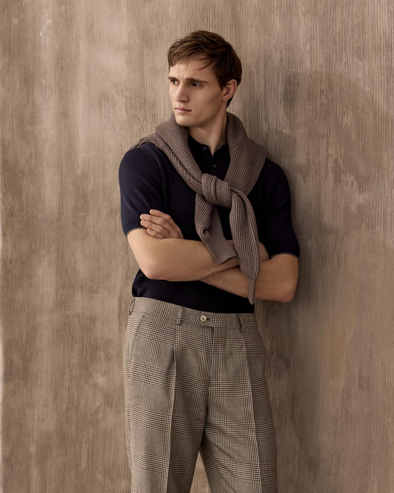 The model is wearing a black wool shirt styled with brown trousers and a brown sweater draped over his shoulders