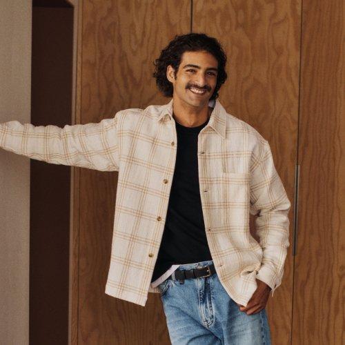 A man wearing a check patterned off-white overshirt with black t-shirt and blue jeans