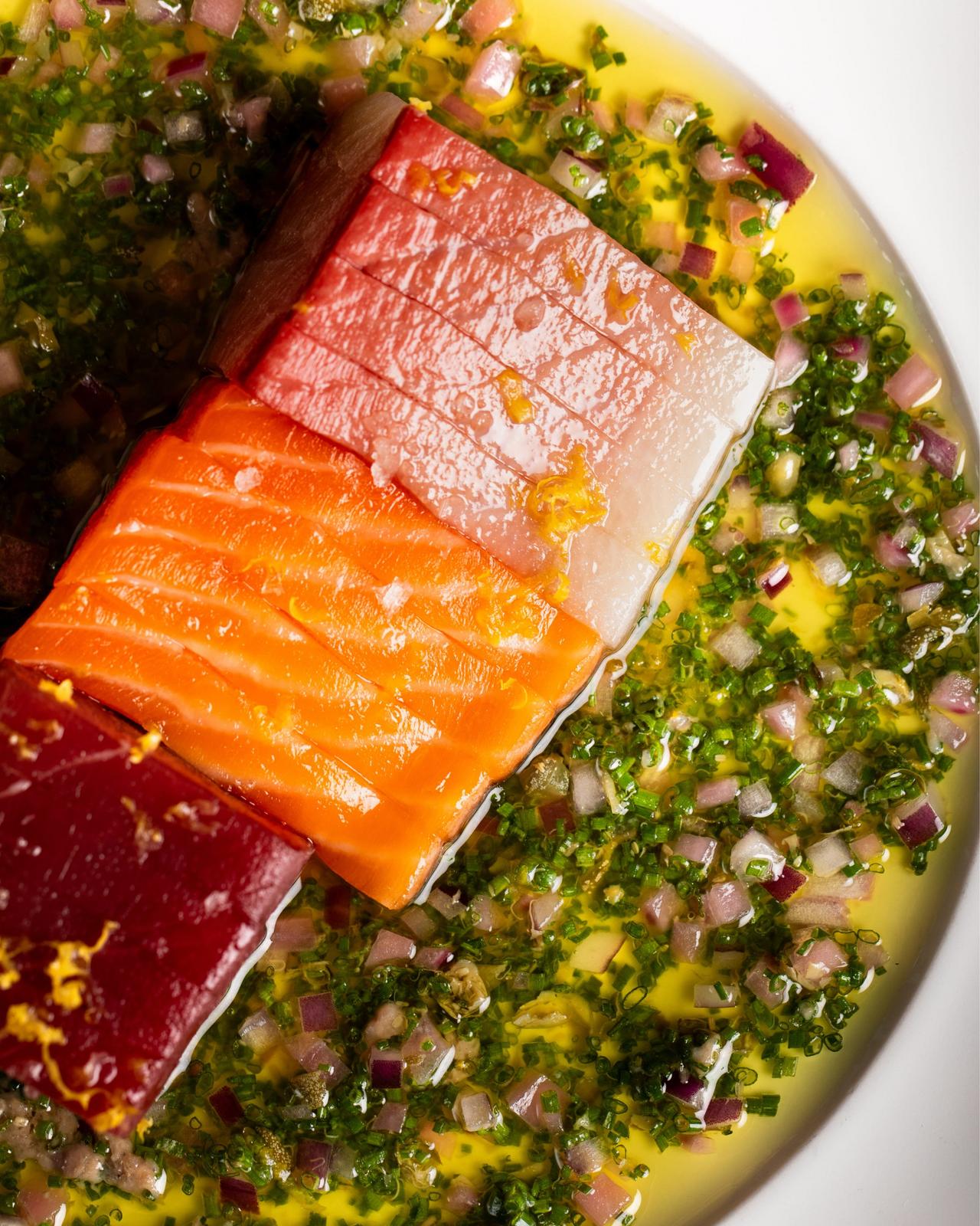 A plate of sashimi served with vibrant salsa verde, featuring slices of salmon, tuna, and mahi 