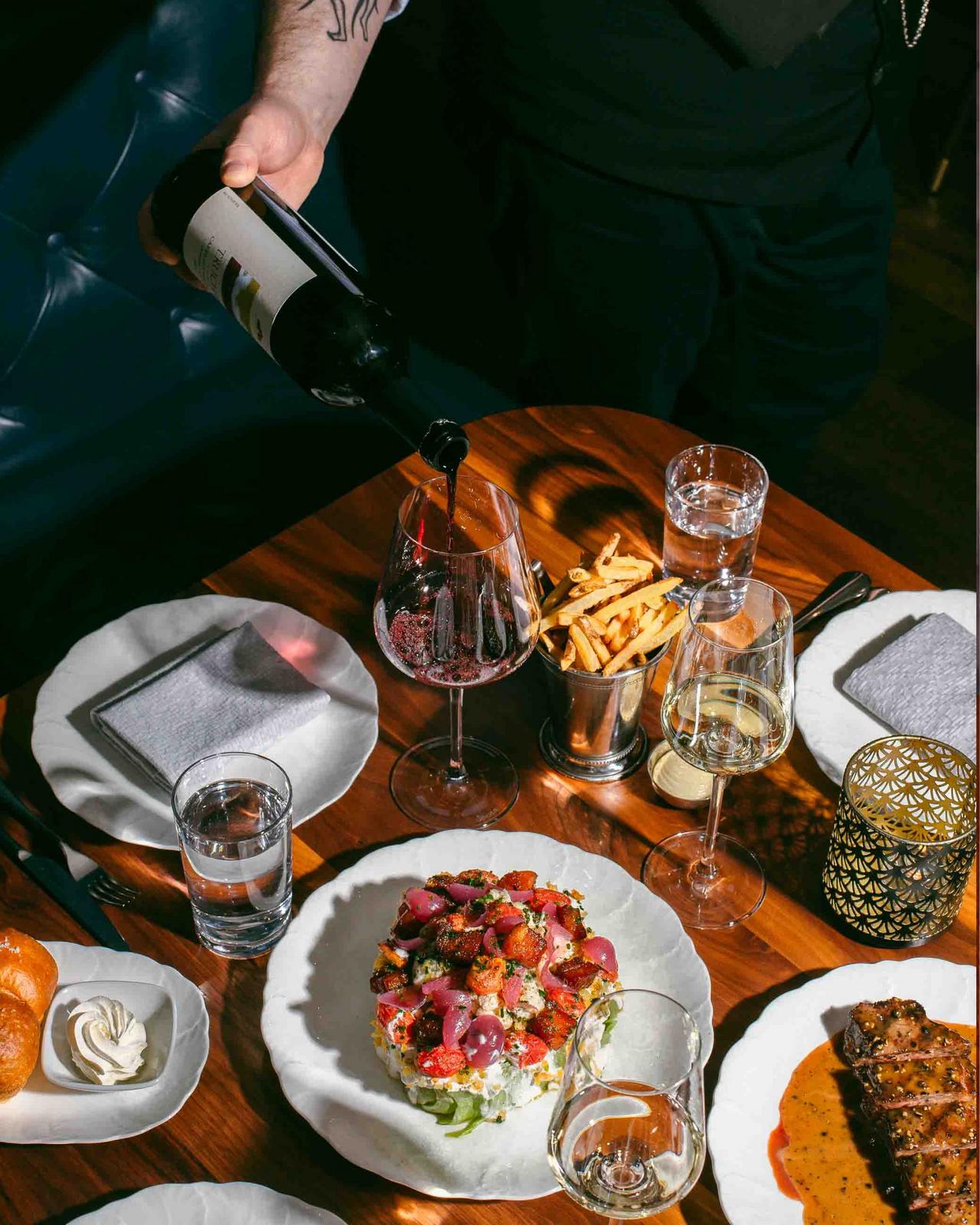 Un plat servi avec un serveur versant du vin rouge dans un verre, accompagné d’un panier de frites dorées