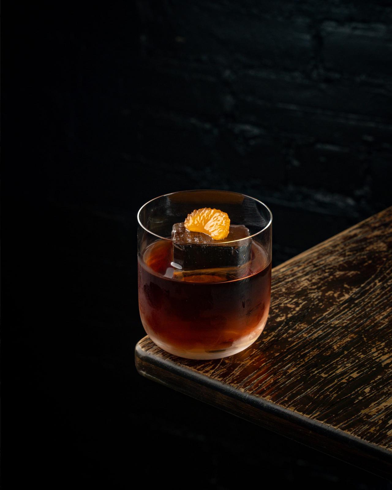 A dark amber cocktail served in a rocks glass, garnished with a large sugar cube and a thin orange slice