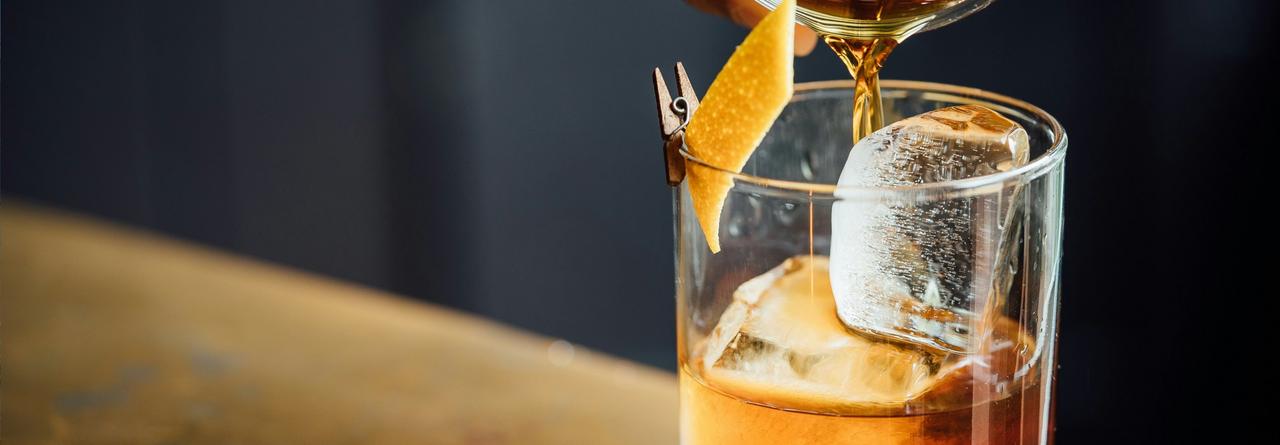 Old Fashioned cocktail, with amber liquid being poured over large ice cubes into a rocks glass