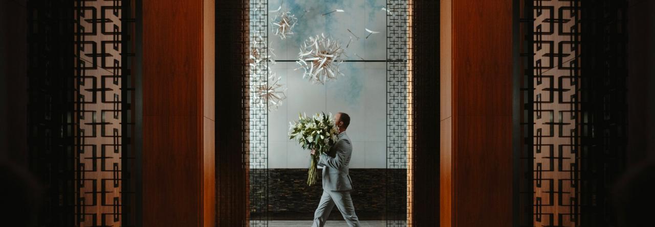 Un homme vêtu d’un complet gris pâle traverse un espace à l’architecture moderne en tenant un grand bouquet de fleurs blanches