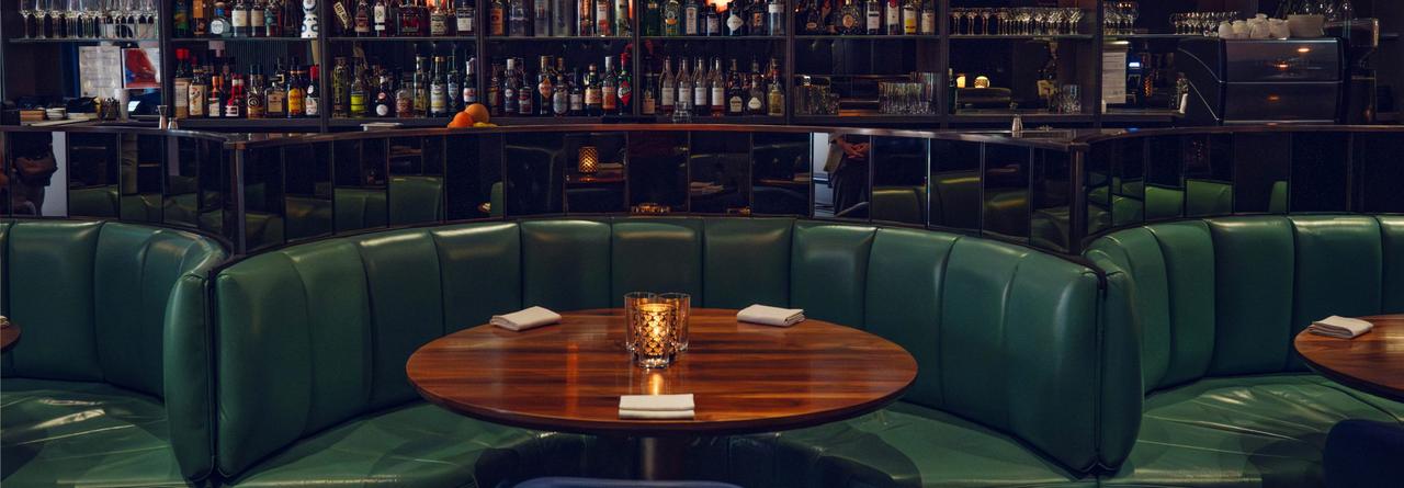Alobar Yorkville, featuring plush green leather booths, a polished wooden table, and a backdrop of an extensive liquor display