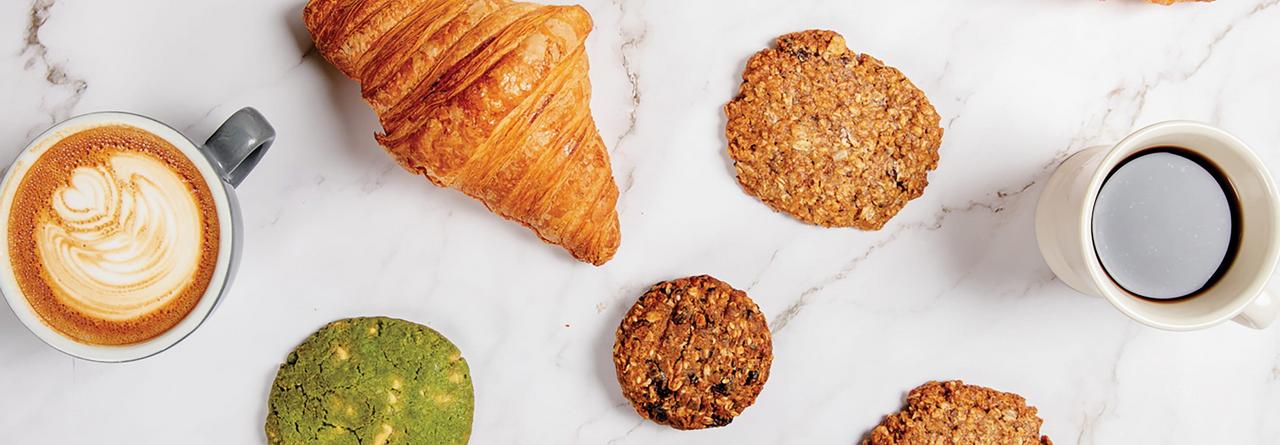 Des pâtisseries et des boissons, comprenant un croissant, des biscuits à l’avoine, un biscuit au matcha et deux tasses de café