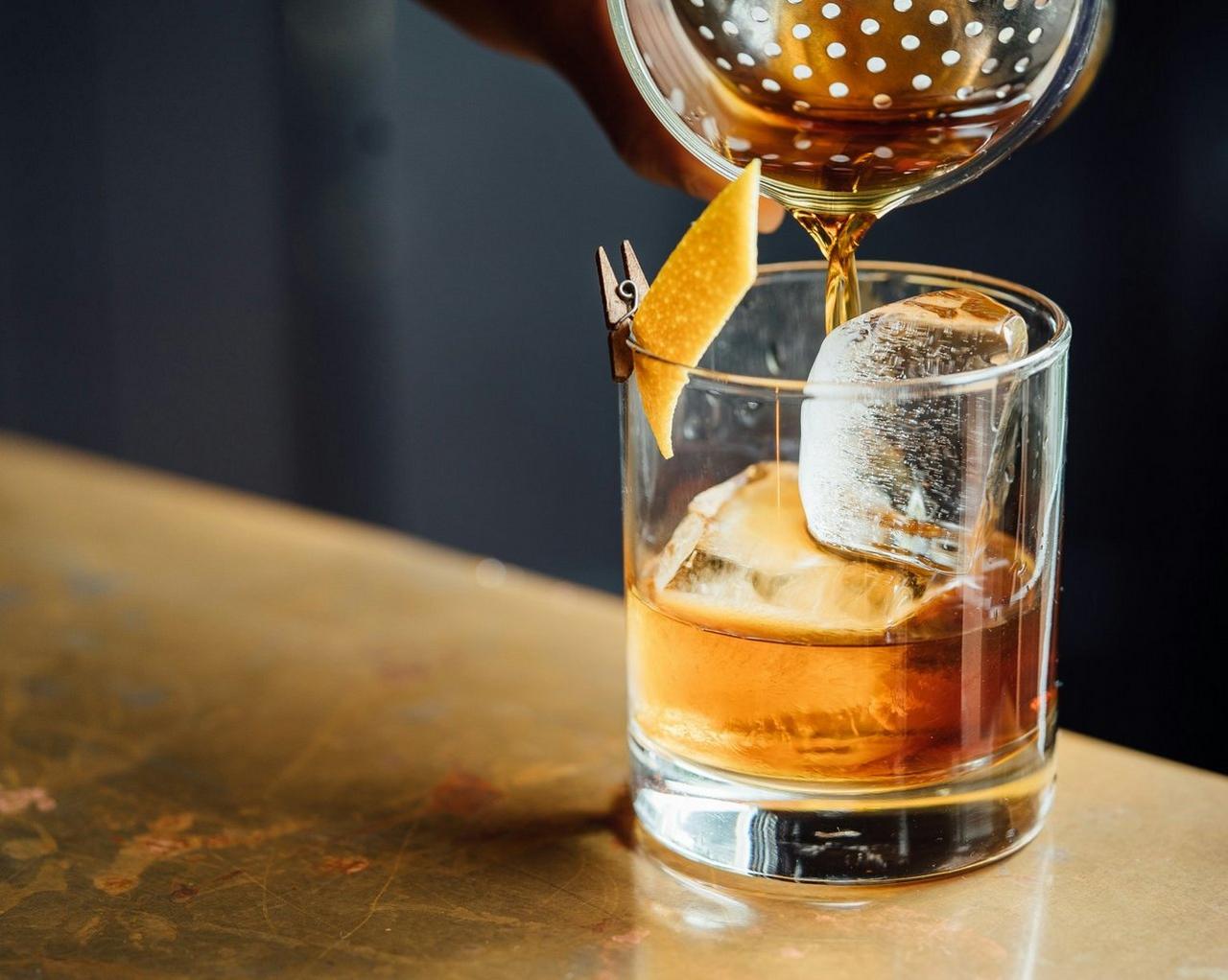 Old Fashioned cocktail, with amber liquid being poured over large ice cubes into a rocks glass