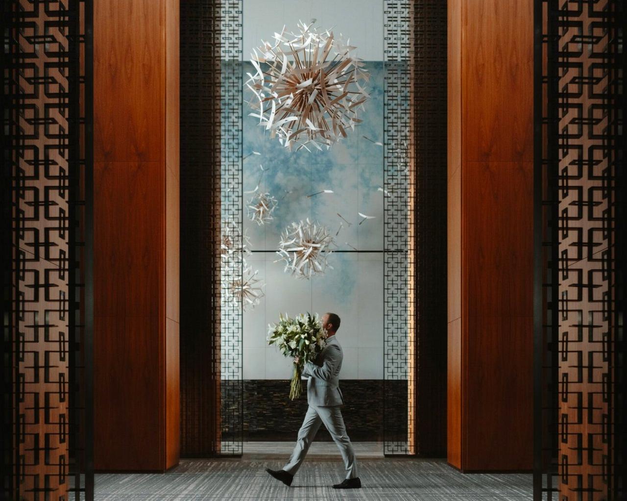 A man in a light gray suit walking through a modern architectural space while holding a large bouquet of white flowers