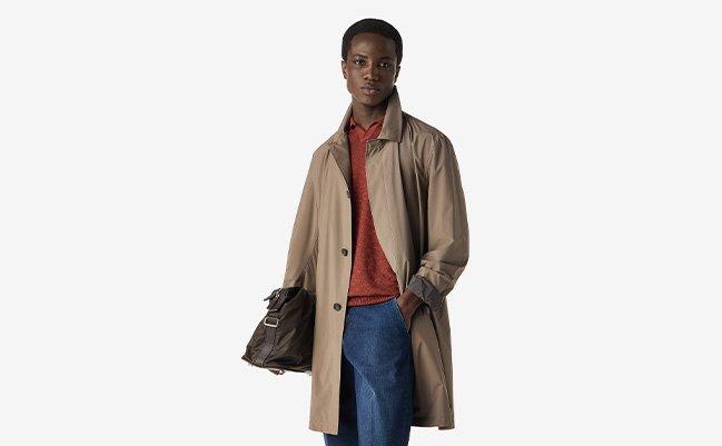 A model wearing a tan lightweight jacket, rust colored polo, styled with denim and a leather bag